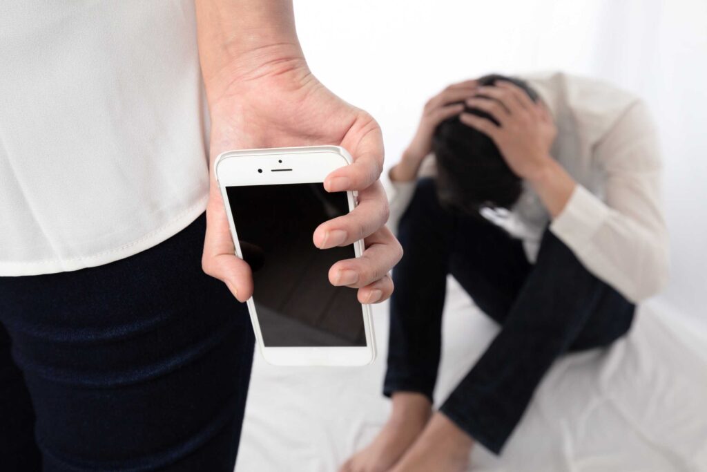 Close-up of a person holding a white smartphone at hip level; in the background, another person grasping their head in distress.