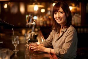 Smiling woman seated at a dimly lit bar, holding a glass of whiskey in front of her.