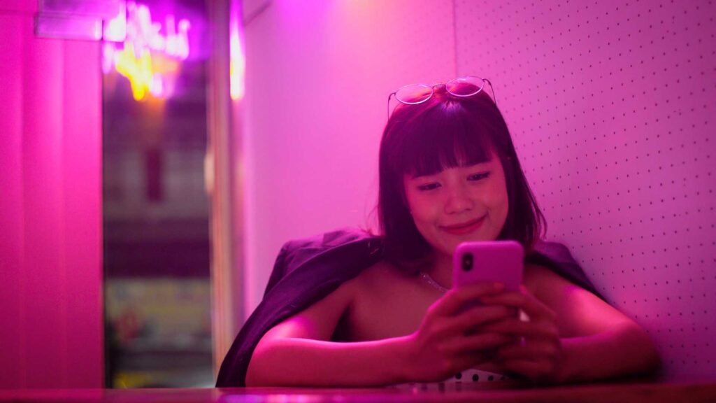 Young woman seated in a pink neon-lit booth, looking at a pink smartphone with sunglasses on her head.