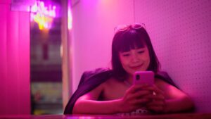 Young woman seated in a pink neon-lit booth, looking at a pink smartphone with sunglasses on her head.